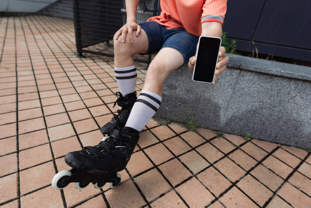 Обрезанный вид человека в роликах и повседневной одежде, показывающий сотовый телефон с пустым экраном на открытом воздухе  - Фото, изображение