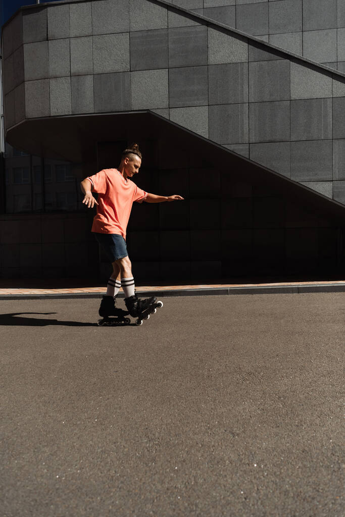 Zijaanzicht van rolschaatser in shorts en t-shirt rijden op asfalt in de buurt van gebouw buiten  - Foto, afbeelding
