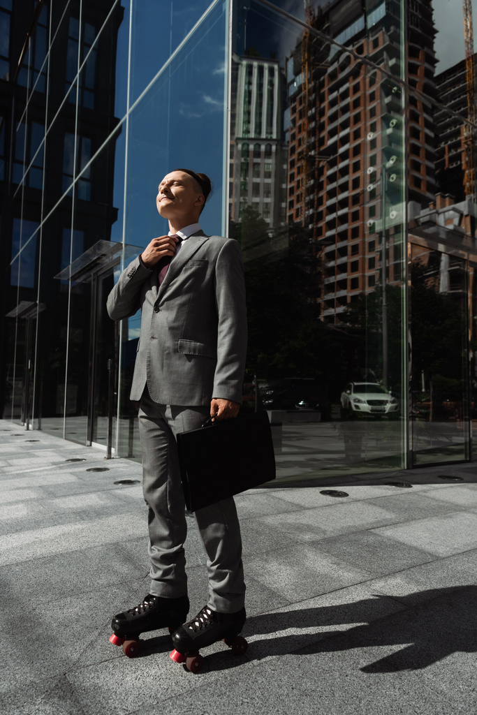 πλήρες μήκος του επιχειρηματία σε γκρι κοστούμι και πατίνια κρατώντας χαρτοφύλακα και στερέωση γραβάτα σε αστικό δρόμο κοντά στο σύγχρονο κτίριο - Φωτογραφία, εικόνα