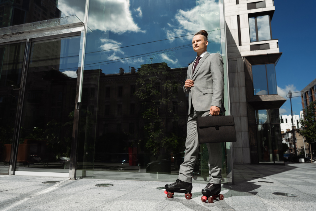 бизнесмен в сером костюме и роликовых коньках стоит с портфелем рядом с современным зданием со стеклянным фасадом - Фото, изображение
