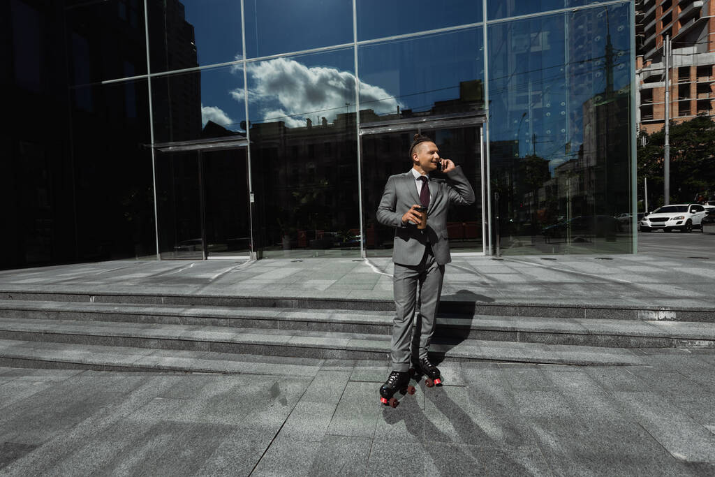 молодой бизнесмен в роликовых коньках разговаривает на смартфоне и держит бумажную чашку возле здания со стеклянным фасадом - Фото, изображение