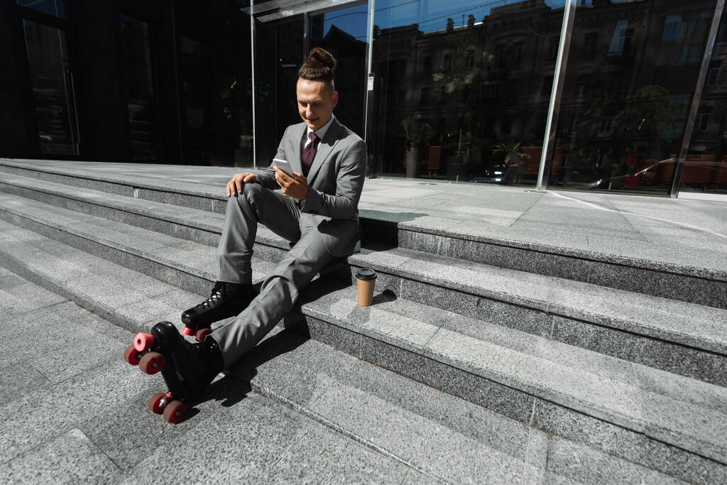 empresario positivo en patines charlando en el teléfono inteligente mientras está sentado cerca de la taza de papel en las escaleras  - Foto, imagen