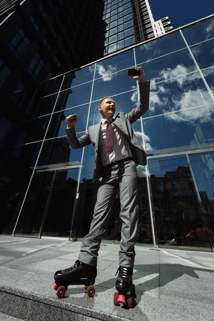 Низкий угол обзора роликового конька в деловом костюме, держащего смартфон и показывающего жест успеха рядом с современным зданием - Фото, изображение