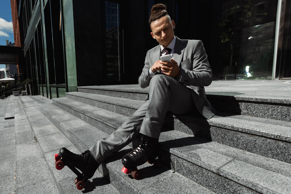 hombre de negocios en patines y auriculares inalámbricos charlando en el teléfono móvil mientras está sentado en las escaleras al aire libre - Foto, imagen