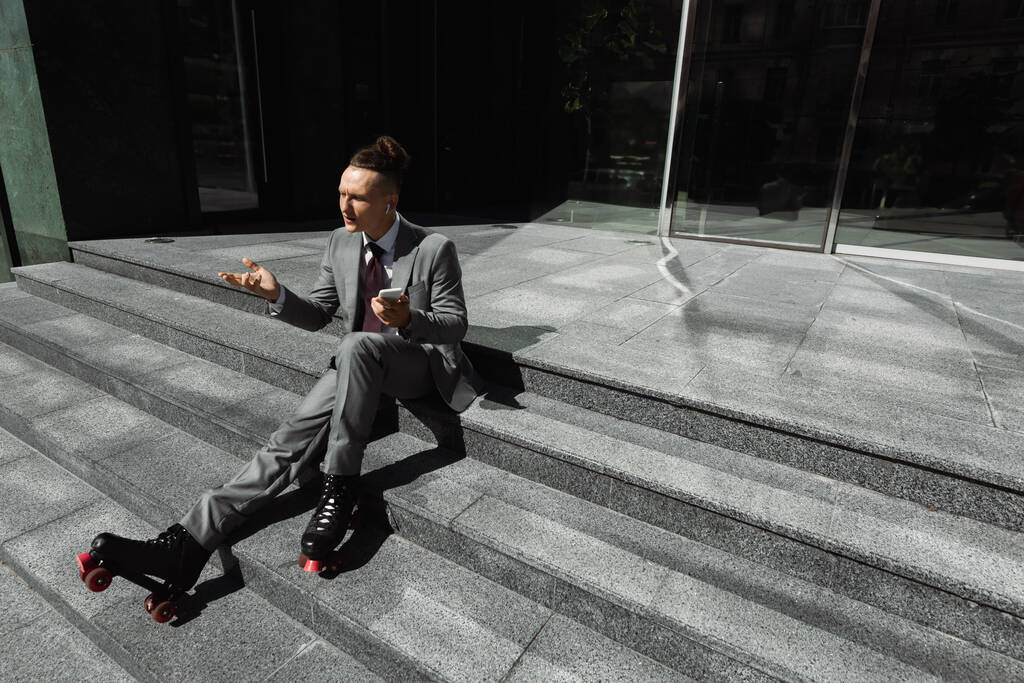 zakenman in pak en rolschaatsen zitten op de trap met mobiele telefoon en tonen schouderophalen gebaar - Foto, afbeelding