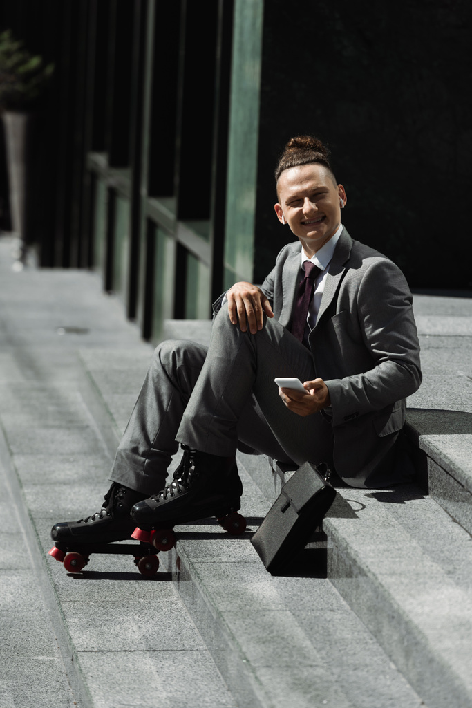 cheerful businessman in roller skates and wireless earphones holding cellphone and looking at camera while sitting on stairs outdoors - Photo, Image