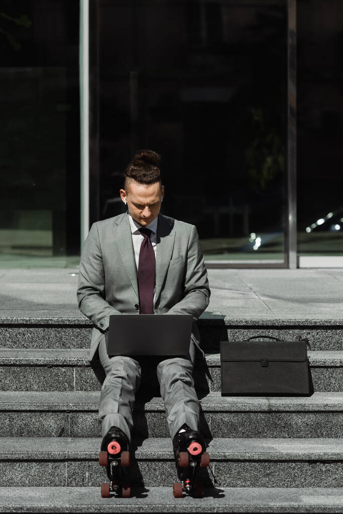 стильний бізнесмен у формальному одязі та роликових ковзанах, що друкуються на ноутбуці, сидячи на сходах біля портфеля
 - Фото, зображення