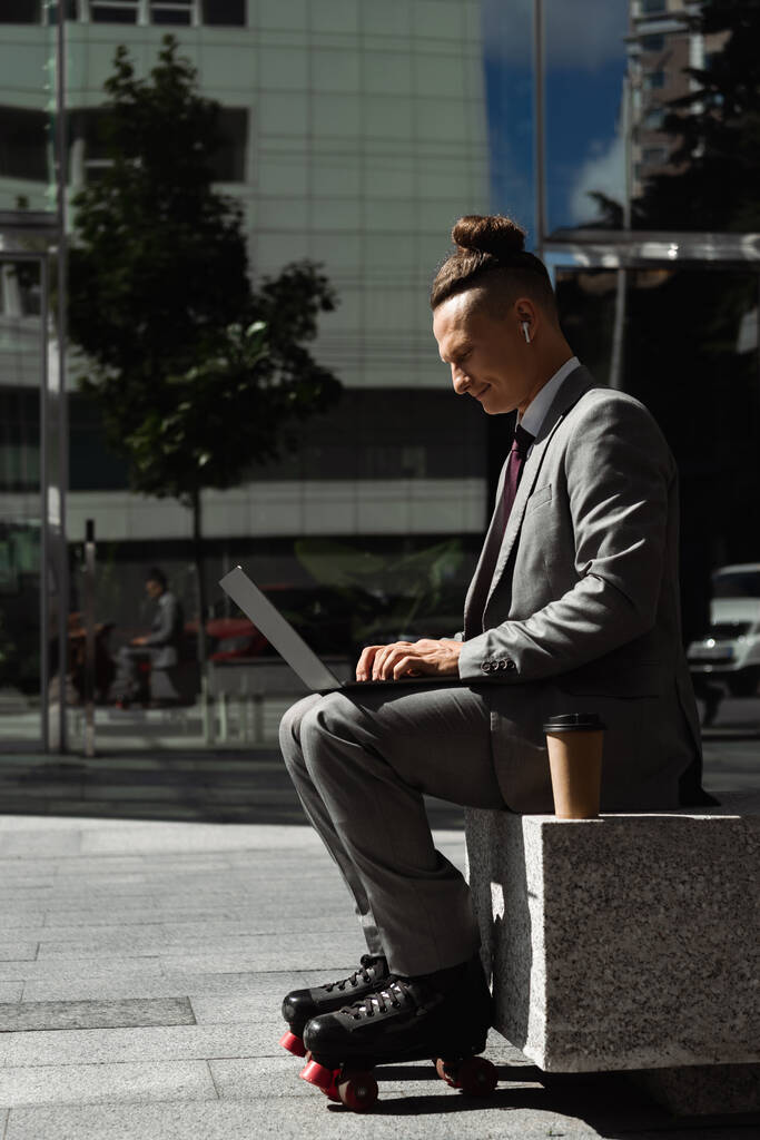 side view of roller skater in grey suit and wireless earphone typing on laptop near coffee to go on urban street - Photo, Image