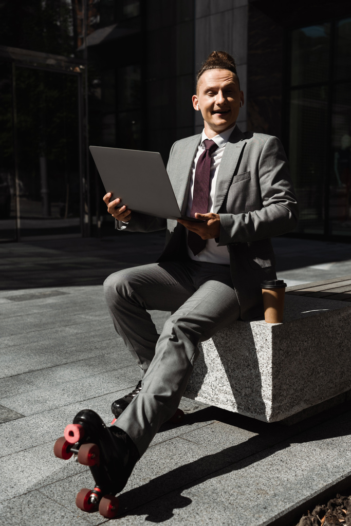 веселый бизнесмен в костюмах и роликовых коньках держит ноутбук и смотрит в камеру на городской улице - Фото, изображение