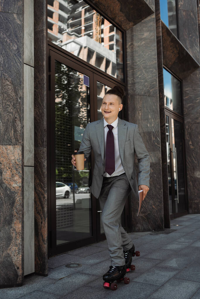 счастливый человек в деловом костюме катается на роликах с бумажной чашкой и папкой на городской улице  - Фото, изображение