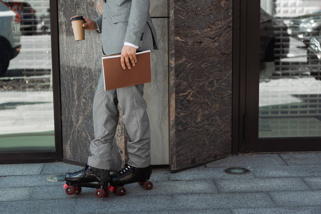 обрізаний вид роликового ковзаняра в костюмі, що стоїть з виносним напоєм і текою біля будівлі в місті
 - Фото, зображення
