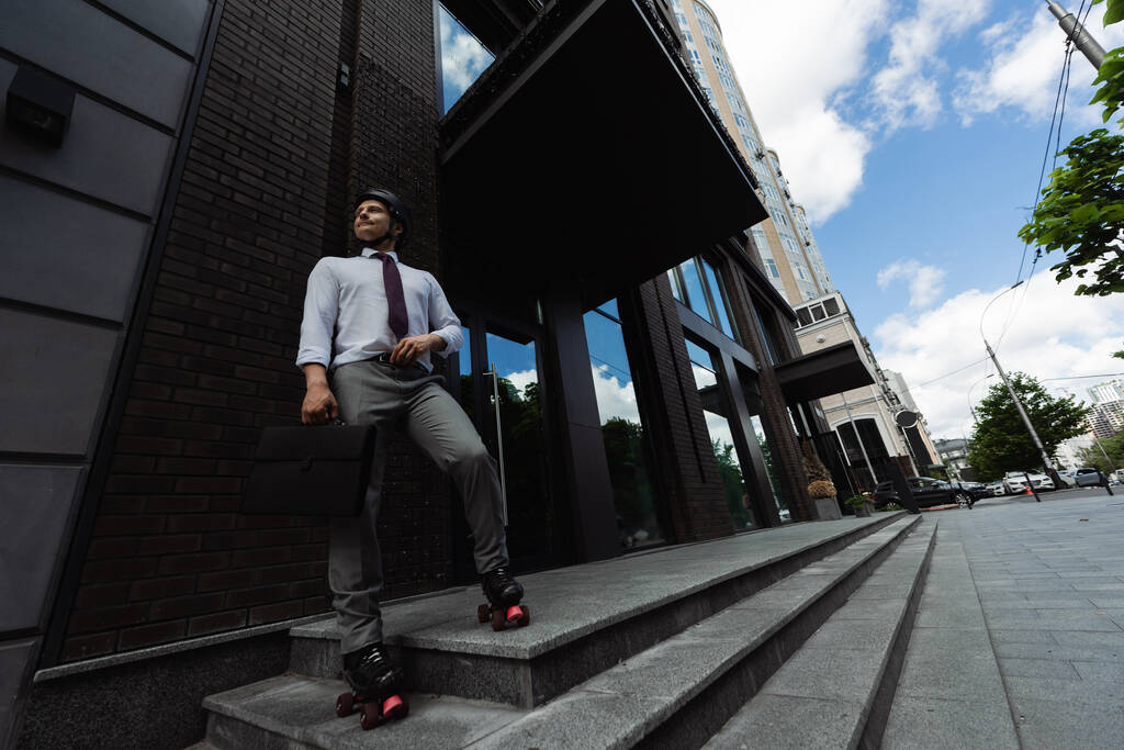 longitud completa del hombre en desgaste formal y patines con maletín y mirando hacia la calle urbana - Foto, imagen