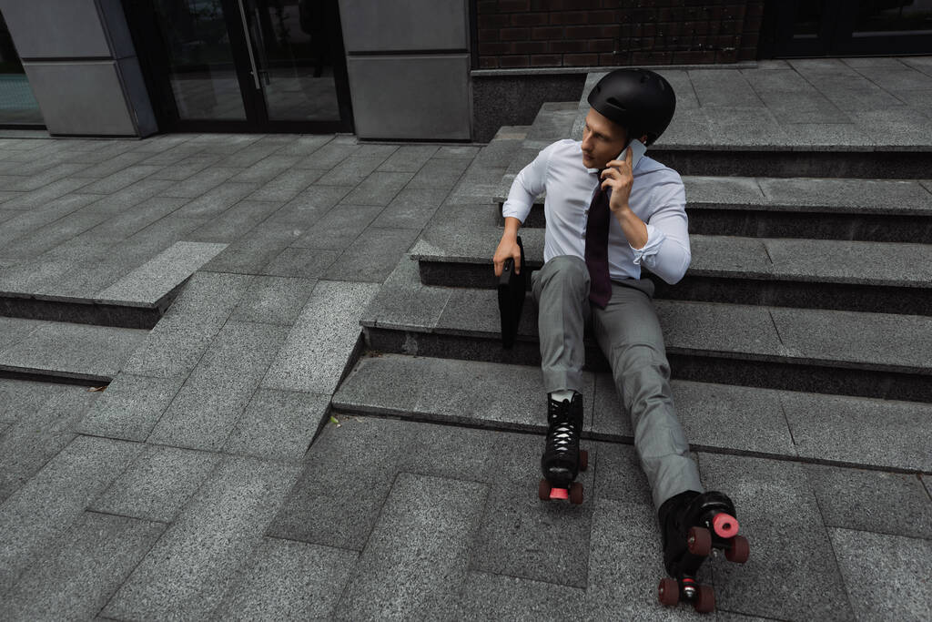 visão de alto ângulo de empresário em capacete e patins falando no smartphone enquanto sentado em escadas ao ar livre - Foto, Imagem