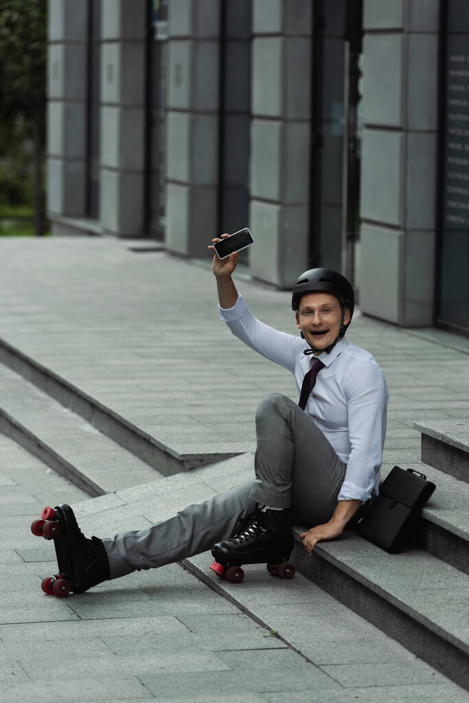 збуджений бізнесмен в шоломі і роликах тримає смартфон з порожнім екраном, сидячи на сходах на відкритому повітрі
 - Фото, зображення