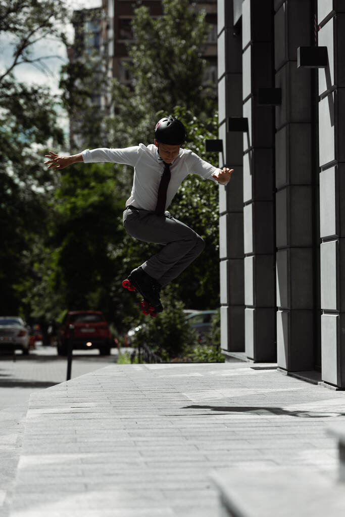 comprimento total do homem no desgaste formal e patins com capacete pulando ao fazer truque na rua urbana - Foto, Imagem