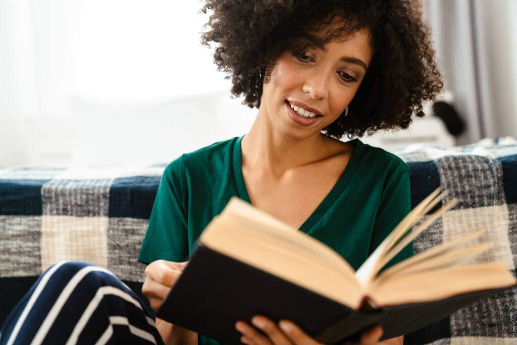 Αφρικανική αμερικανική νεαρή γυναίκα με σγουρά afro χτένισμα χαμογελώντας και διαβάζοντας το βιβλίο στο σπίτι - Φωτογραφία, εικόνα