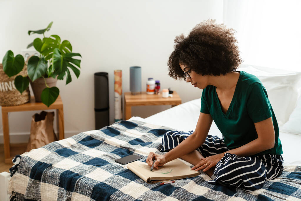 Afričanky americká mladá žena s kudrnatý účes sedí v posteli a kreslení v skicáku doma - Fotografie, Obrázek
