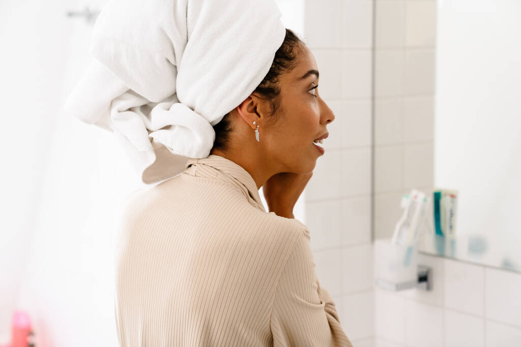 Afroamericana giovane donna con asciugamano sulla testa sorridente e guardando nello specchio al bagno - Foto, immagini