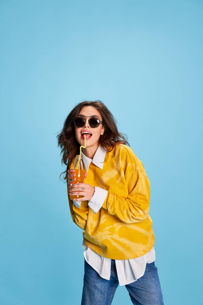 Retrato de una joven hermosa bebiendo jugo de naranja, posando en gafas de sol aisladas sobre fondo azul. Casual. Concepto de juventud, belleza, moda, estilo de vida, emociones, expresión facial. Anuncio - Foto, imagen