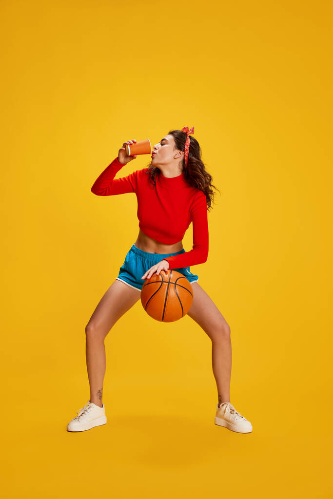 Portrait of young beautiful girl posing with basketball ball and drinking protein isolated over yellow background. Concept of youth, beauty, sport lifestyle, emotions, facial expression. Ad - Photo, Image