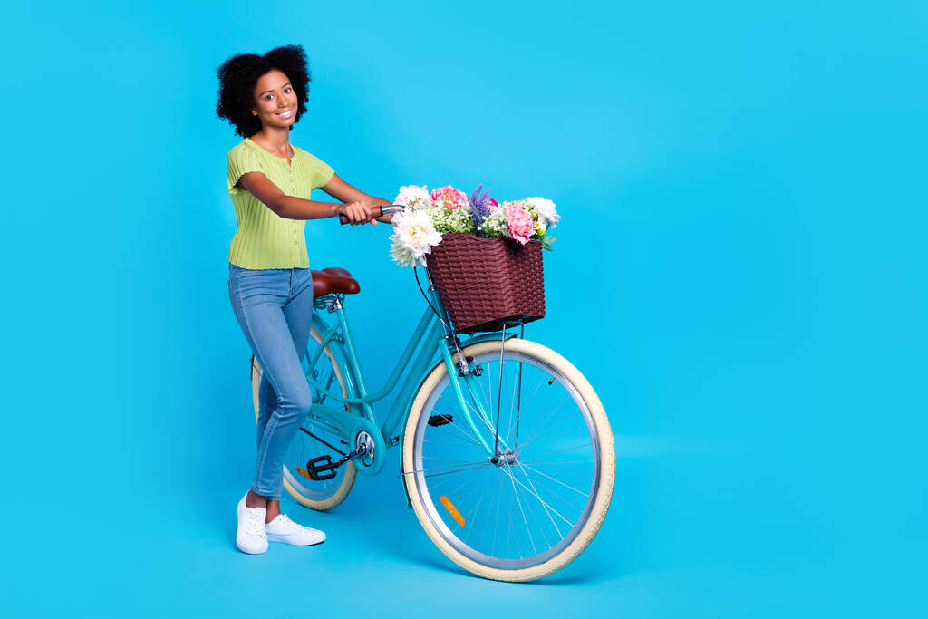 Küçük neşeli, neşeli genç bayanın tam boy fotoğrafı mavi arka planda izole edilmiş çiçek teslimatlı bisiklet sepetini tutuyor.. - Fotoğraf, Görsel