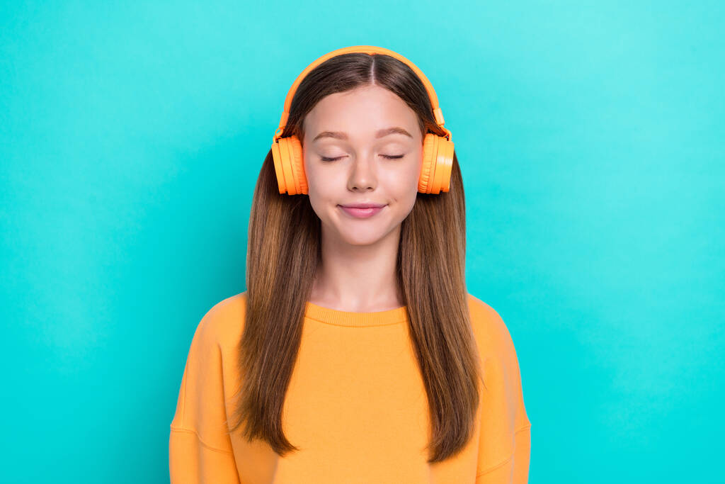 Foto de una joven chica divertida y soñadora que usa auriculares inalámbricos naranjas ojos cerrados escucha su banda sonora de preferencia aislada en el fondo de color aguamarina. - Foto, imagen