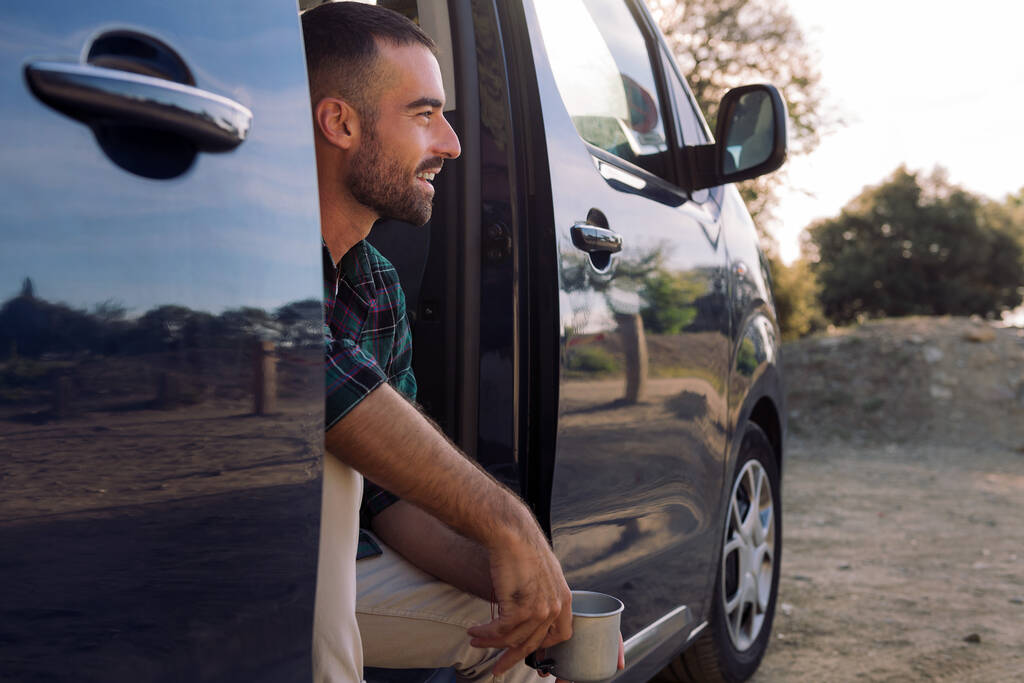 улыбающийся молодой человек, сидящий в дверях своего фургона с кружкой в руке, концепцией свободы и кочевого образа жизни - Фото, изображение