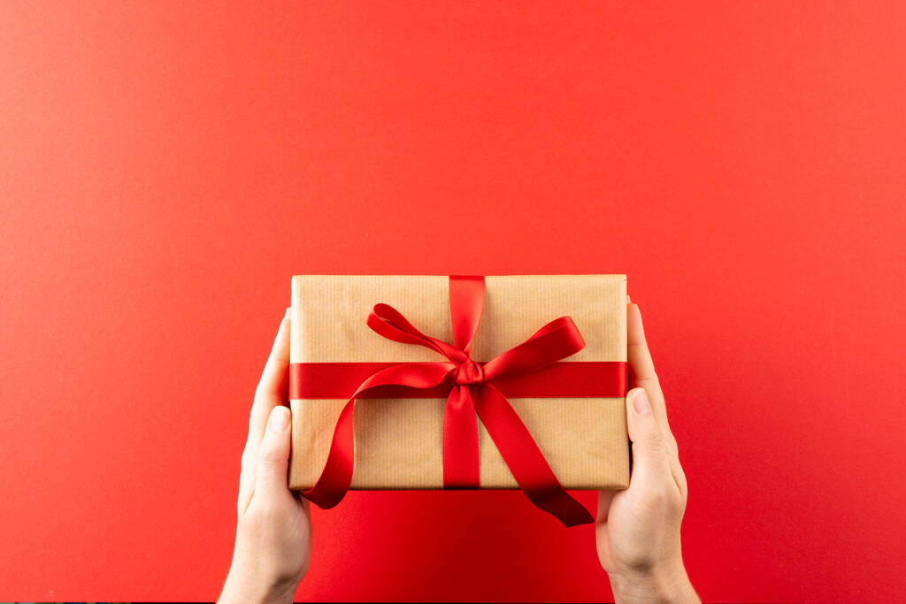 Overhead of hands met cadeau in bruin papier met rood lint, op rode achtergrond met kopieerruimte. Valentijnsdag, liefde, romantiek en feest concept. - Foto, afbeelding