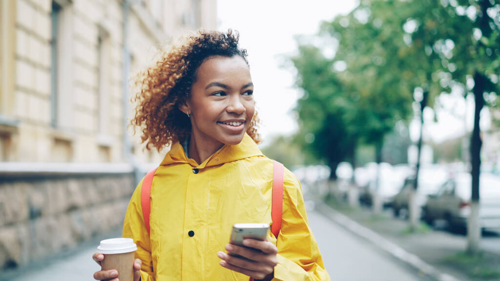 Boldog afrikai-amerikai nő használ smartphone képernyő megérintésével, és mosolyogva, séta kint gyönyörű város megy a kávé. Modern életmód, az emberek és a kommunikáció fogalma. - Fotó, kép