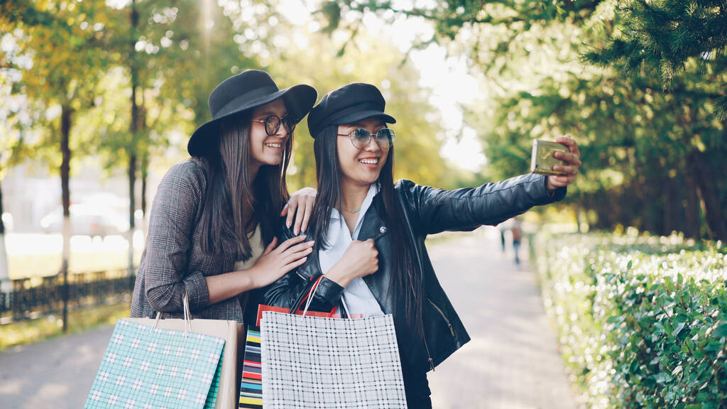 Gelukkig jonge vrouwelijke studenten zijn het maken van online video-oproep met slimme telefoon over winkelen weergegeven: tassen en lachen staande in de straat op herfstdag. - Foto, afbeelding
