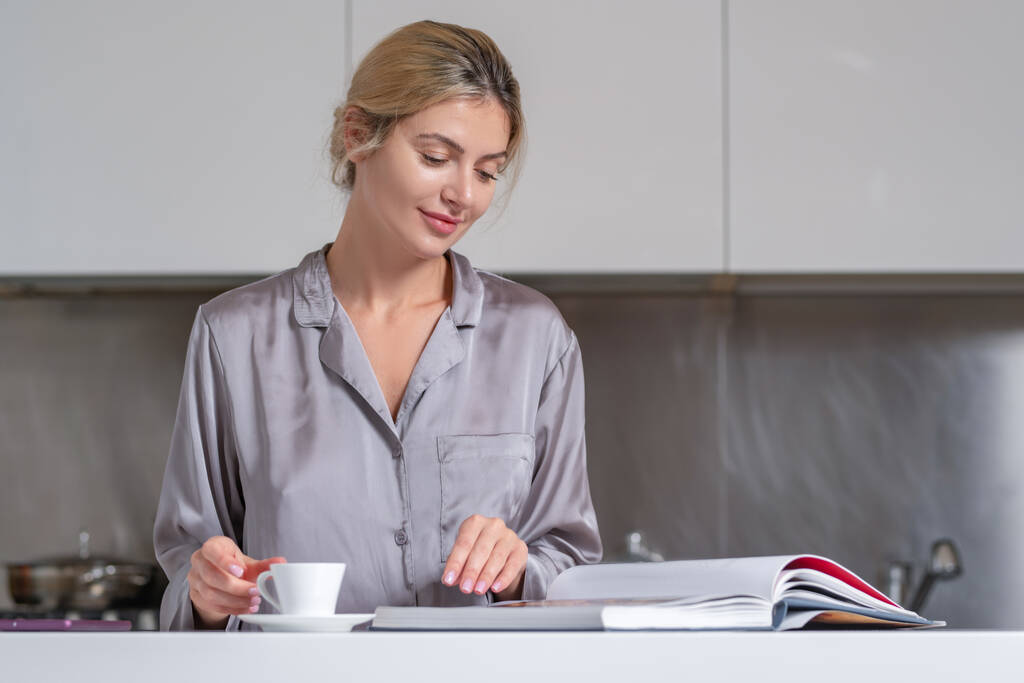 Sexy woman drink coffee and reading book or magazine at home in morning. Beautiful sensual female housewife have breakfast in sexy pajama at kitchen - Photo, Image