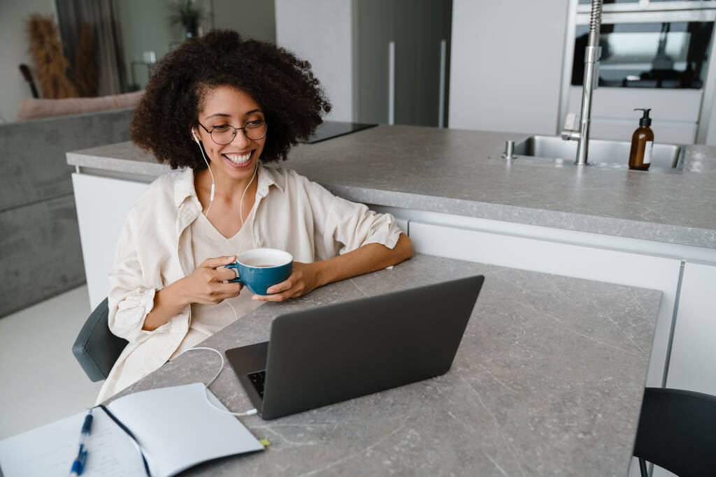 Black woman in earphones drinking coffee while working with laptop at home - Photo, Image
