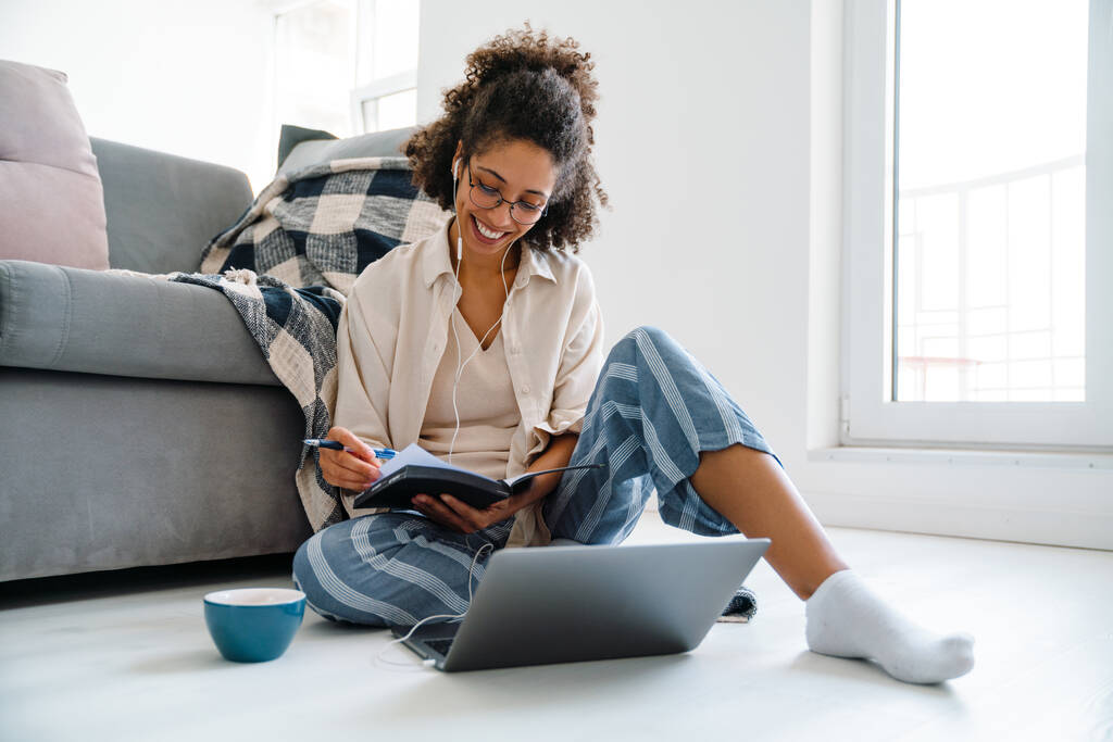 Чорна жінка в навушниках працює з ноутбуком, сидячи на підлозі вдома
 - Фото, зображення