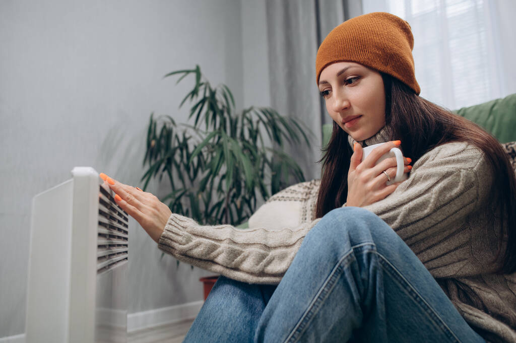 Een warm geklede jonge vrouw warmt zich op bij een elektrische radiator en drinkt warme thee.. - Foto, afbeelding