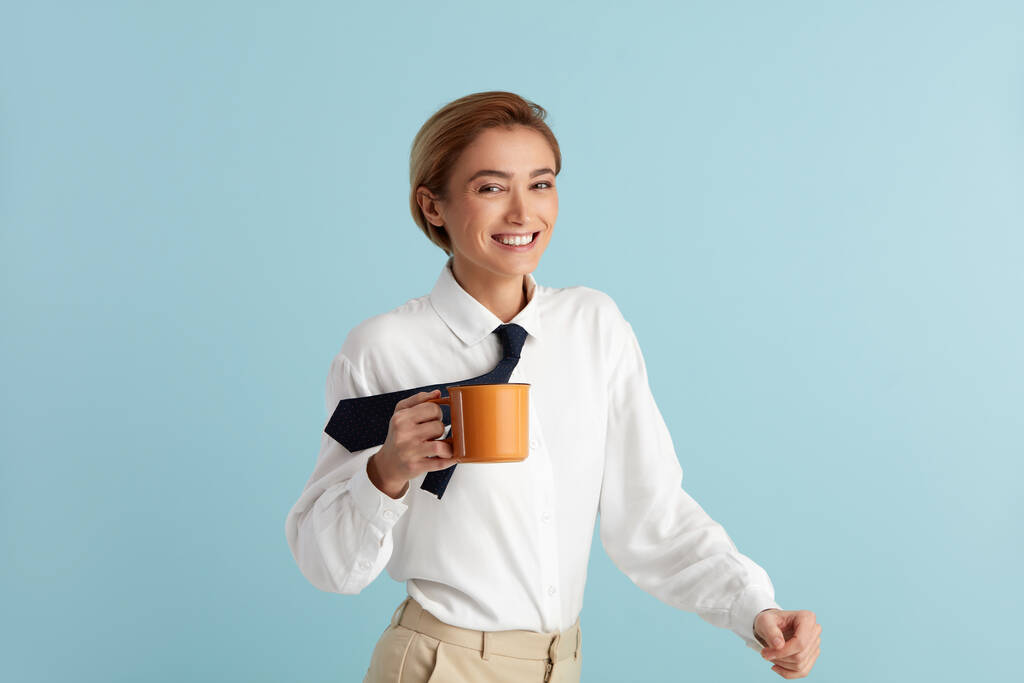 Улыбающаяся деловая женщина, держащая кофе. Портрет дружелюбной молодой женщины, пьющей кофе, держащей напиток в чашке и улыбающейся. Крытая студия, снятая на голубом фоне  - Фото, изображение