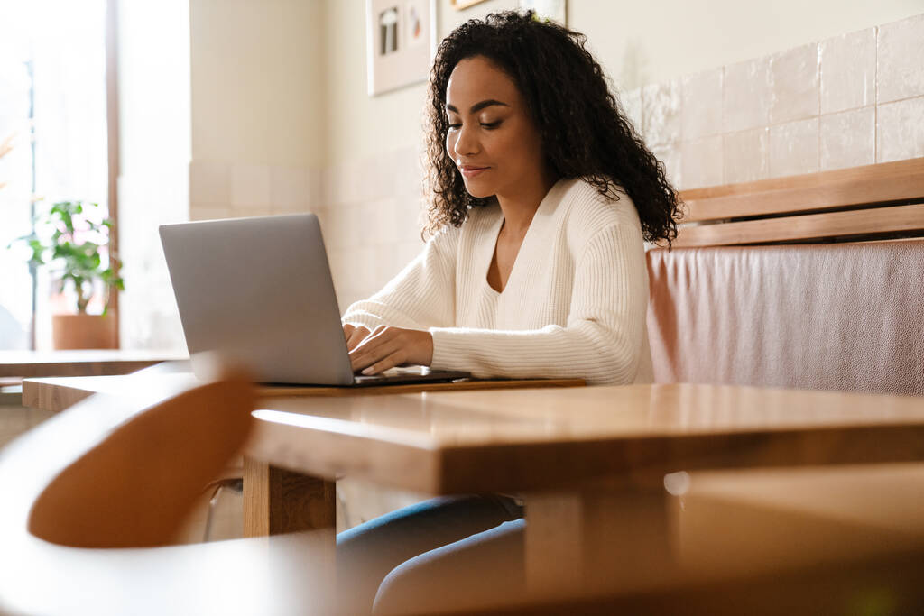 Mujer negra joven que trabaja con el ordenador portátil mientras está sentado en la cafetería en el interior - Foto, imagen
