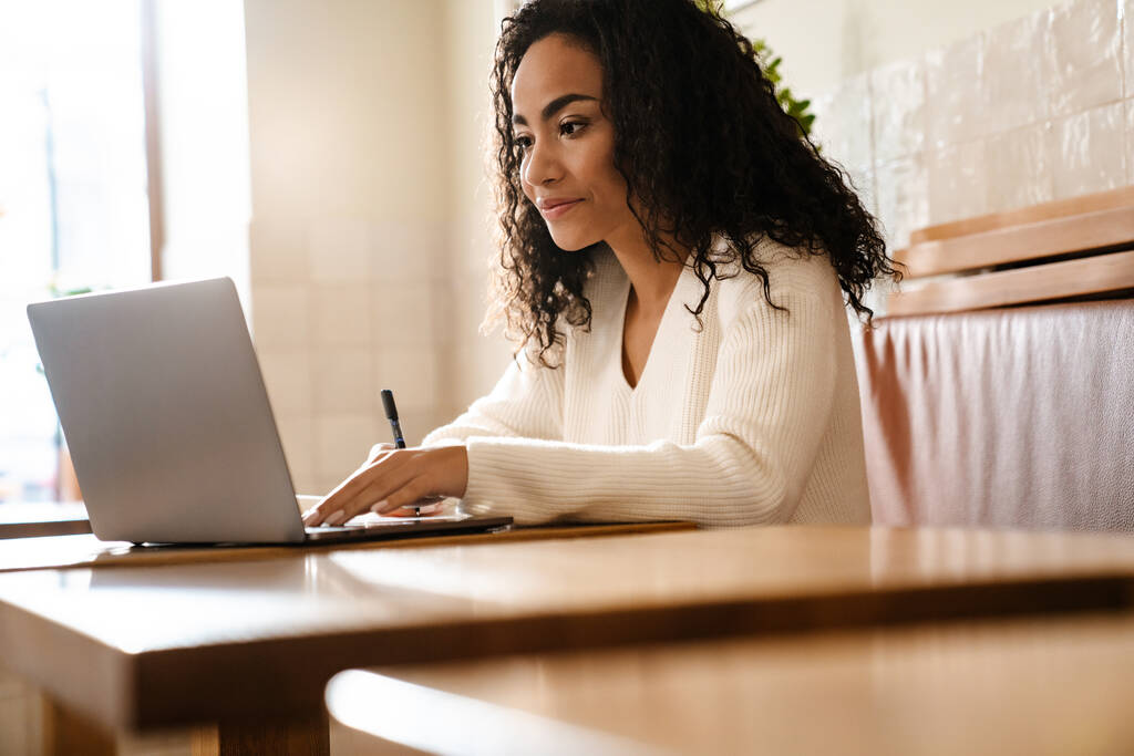 Nuori musta nainen kirjoittaa muistiinpanoja työskennellessään kannettavan tietokoneen kanssa kahvilassa sisätiloissa - Valokuva, kuva