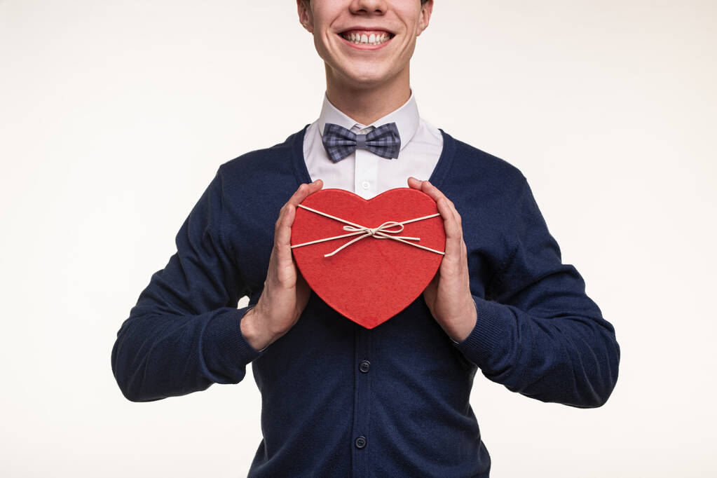 Растение веселый молодой романтичный человек в белой рубашке и галстук-бабочка счастливо улыбаясь, демонстрируя красный сердце форме подарочной коробки на белом фоне - Фото, изображение