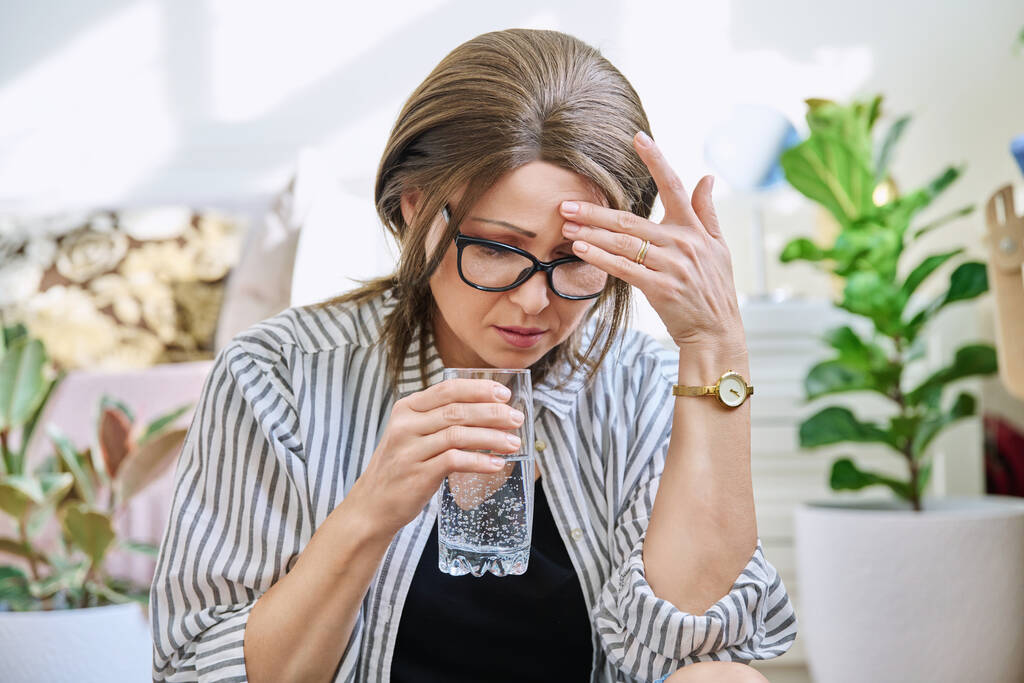 Mujer madura que sufre de dolor de cabeza, en casa con un vaso de agua. Salud, estrés, depresión, dolor, migraña, problemas de edad avanzada - Foto, Imagen