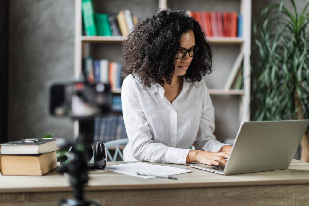 Piękna wielonarodowa kobieta w białej koszuli siedząca przy biurku, pracująca na laptopie i nagrywająca wideo na cyfrowym smartfonie. Koncepcja wolnego strzelca i nowoczesnej technologii. - Zdjęcie, obraz