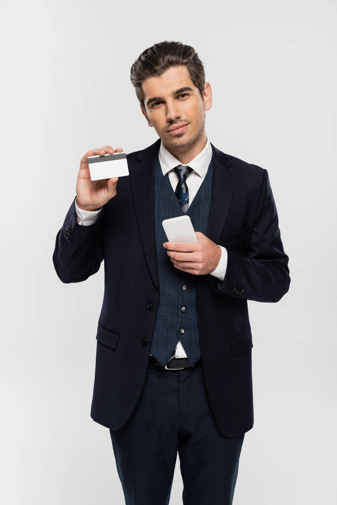 mladý podnikatel v obleku drží smartphone a kreditní karty izolované na šedé  - Fotografie, Obrázek