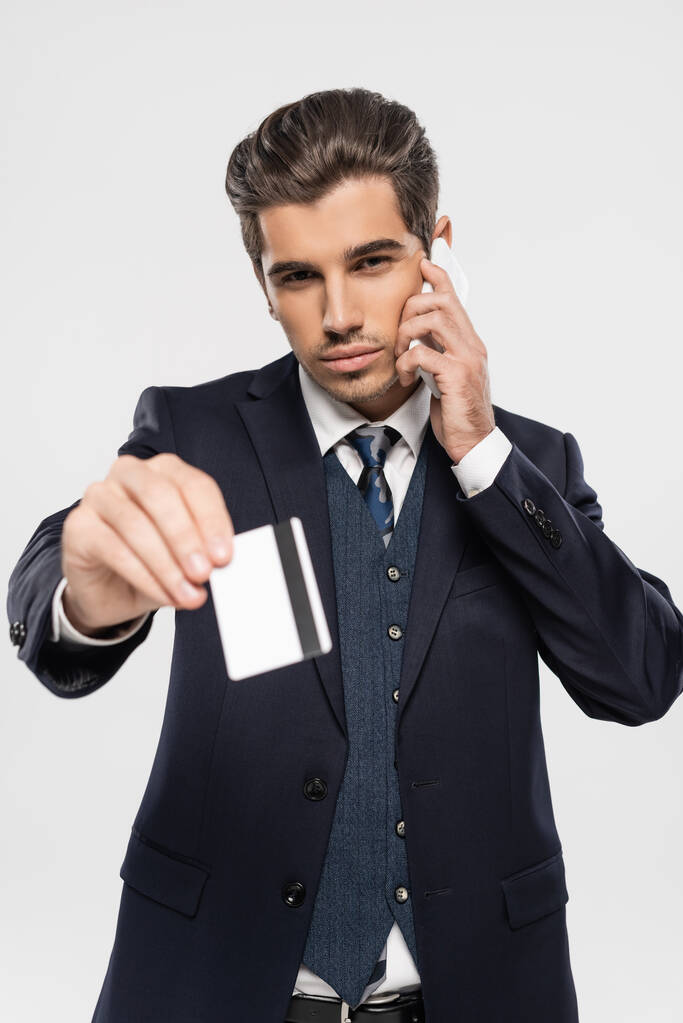 confident businessman in suit talking on smartphone and holding credit card isolated on grey  - Photo, Image