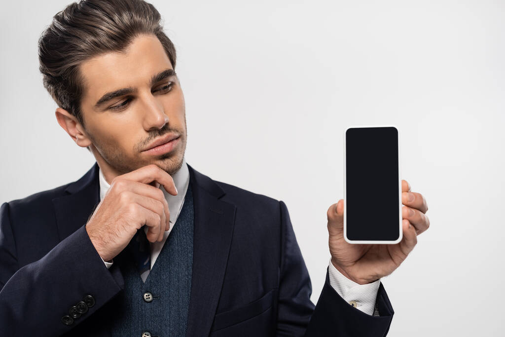 Nachdenklicher Geschäftsmann im Anzug blickt auf Smartphone mit leerem Bildschirm isoliert auf grau  - Foto, Bild