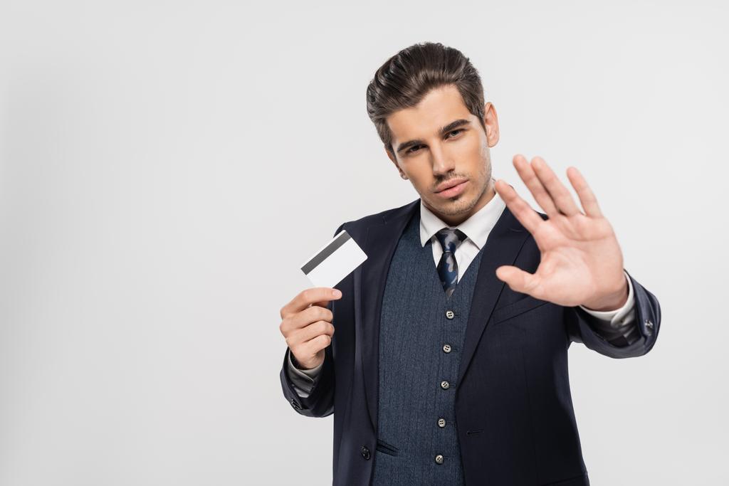 erfolgreicher Geschäftsmann im Anzug hält Kreditkarte und zeigt Stopp-Geste isoliert auf grau - Foto, Bild