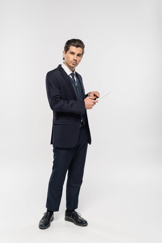 intera lunghezza del giovane uomo d'affari in giacca e cravatta con tablet digitale su grigio  - Foto, immagini