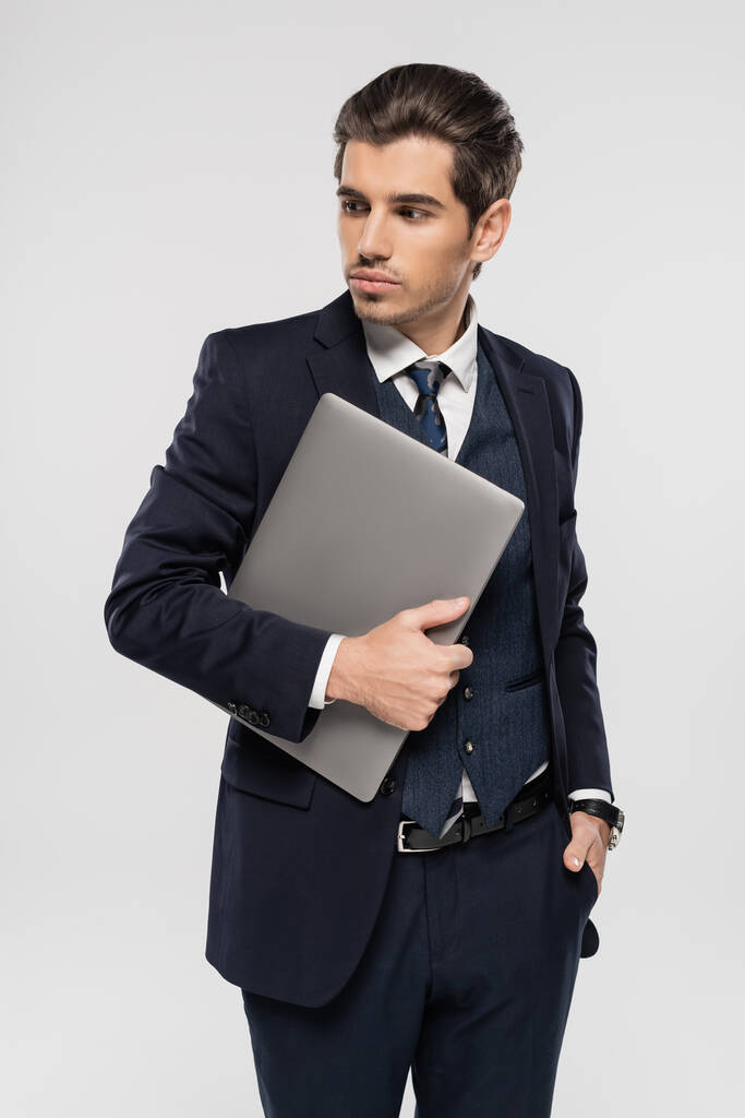 młody biznesmen w garniturze trzyma laptopa i stoi z ręką w kieszeni odizolowany na szaro - Zdjęcie, obraz