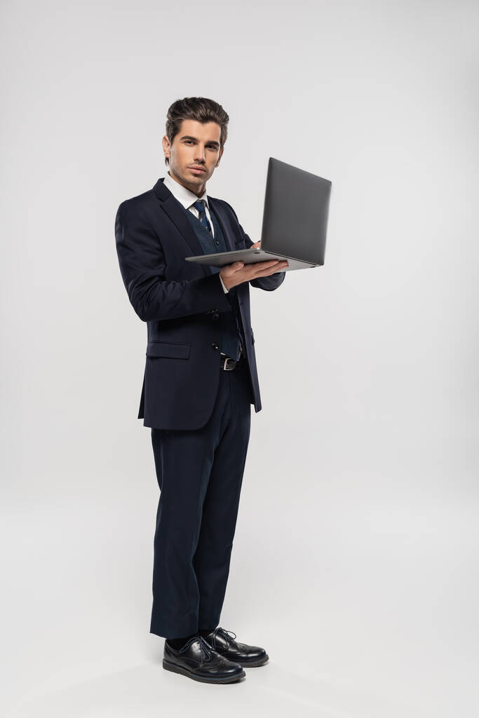 pełna długość pewny biznesmen w garniturze za pomocą laptopa podczas pracy zdalnie na szary - Zdjęcie, obraz