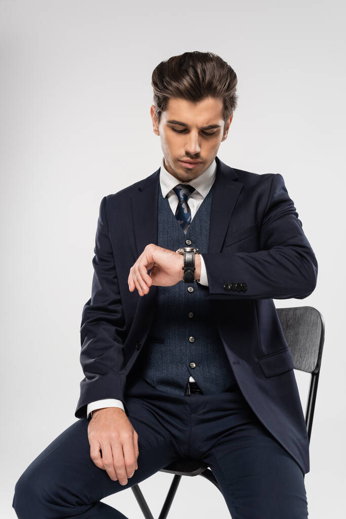 νεαρός επιχειρηματίας με επίσημη ένδυση κάθεται στην καρέκλα, ενώ κοιτάζοντας ρολόι χειρός απομονώνονται σε γκρι - Φωτογραφία, εικόνα