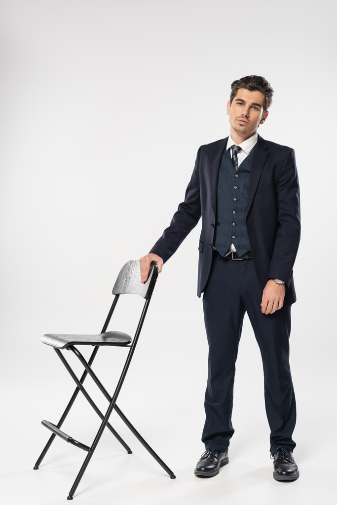longitud completa de joven hombre de negocios en ropa formal de pie cerca de la silla mientras posa en gris - Foto, Imagen