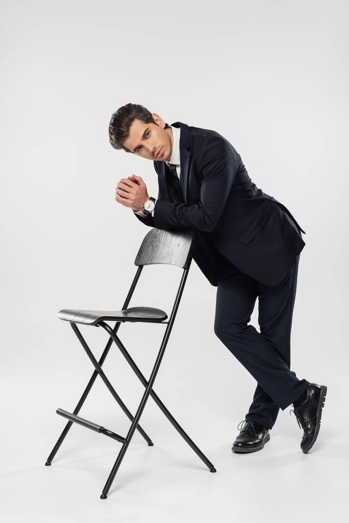pełna długość odnoszącego sukcesy biznesmena w formalnym ubraniu stojącego w pobliżu krzesła na szarym - Zdjęcie, obraz